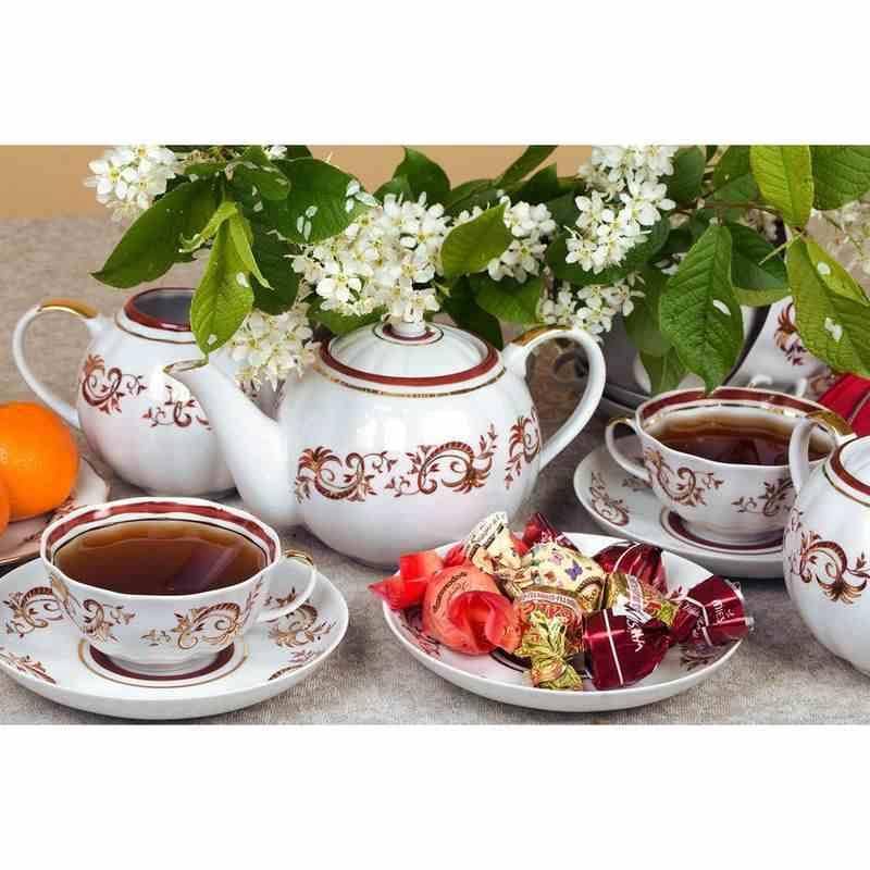 Элегантный чайный сервиз с золотисто-красным узором, чашки, заварник, апельсины и конфеты на фоне цветов